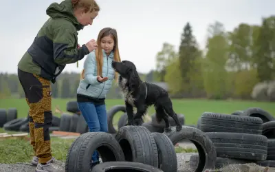En kvinna och en flicka leker med en svart hundvalp bland gamla däck utomhus.