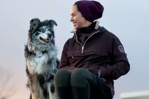 En kvinna ler mot sin border collie med blå ögon. Hunden bär ett halsband med en lysande blå enhet.