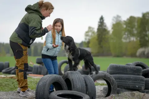 En kvinna och en flicka leker med en svart hundvalp bland gamla däck utomhus.